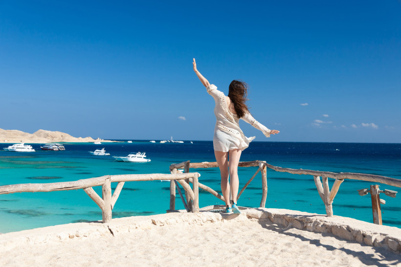Ägypten - Hurghada Eine Frau steht auf einer kleinen Sandterrasse oberhalb des Meeres und blickt in Richtung Wasser. Sie trägt ein weißes, luftiges Strandkleid, ihre Haare wehen im Wind. Vor ihr liegt das azurblaue Meer, gesäumt von mehreren Yachten und Booten, im Hintergru