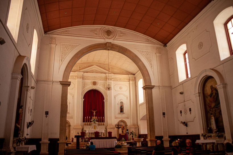 Innenansicht einer katholischen Kirche mit hohen weißen Wänden, großen Rundbogen, klassizistischen Säulen und einem roten Vorhang hinter dem Altar. Der Altarraum ist geschmückt mit Kerzen und Blumen, einige Menschen sitzen in den Bänken
