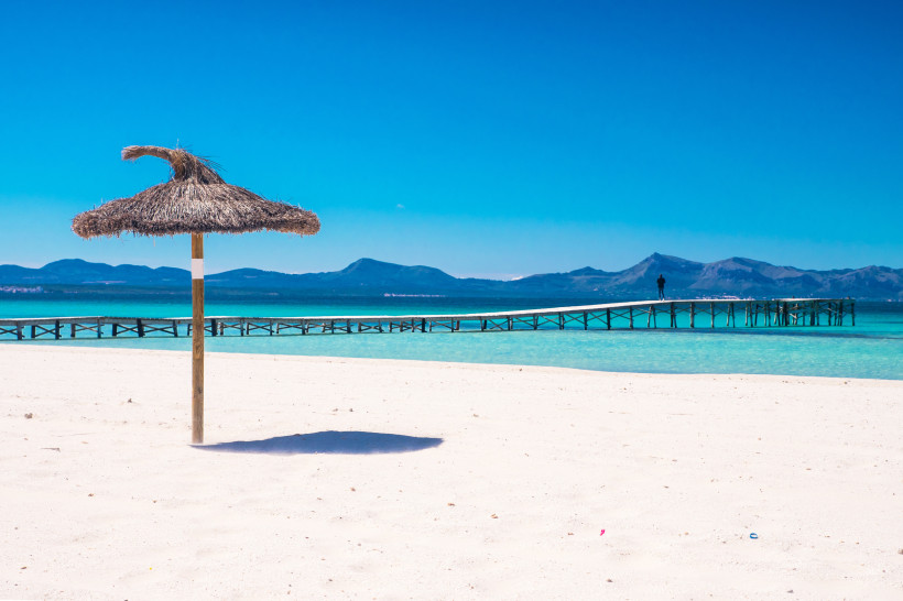 Strohsonnenschirm am Sandstrand der Playa de Muro auf Mallorca, im Hintergrund ein Steg über türkisfarbenes Meer und Berge