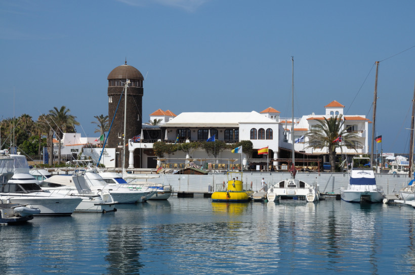 Hafen von Caleta de Fuste mit Yachten am Steg, weißem Gebäude und Leuchtturm