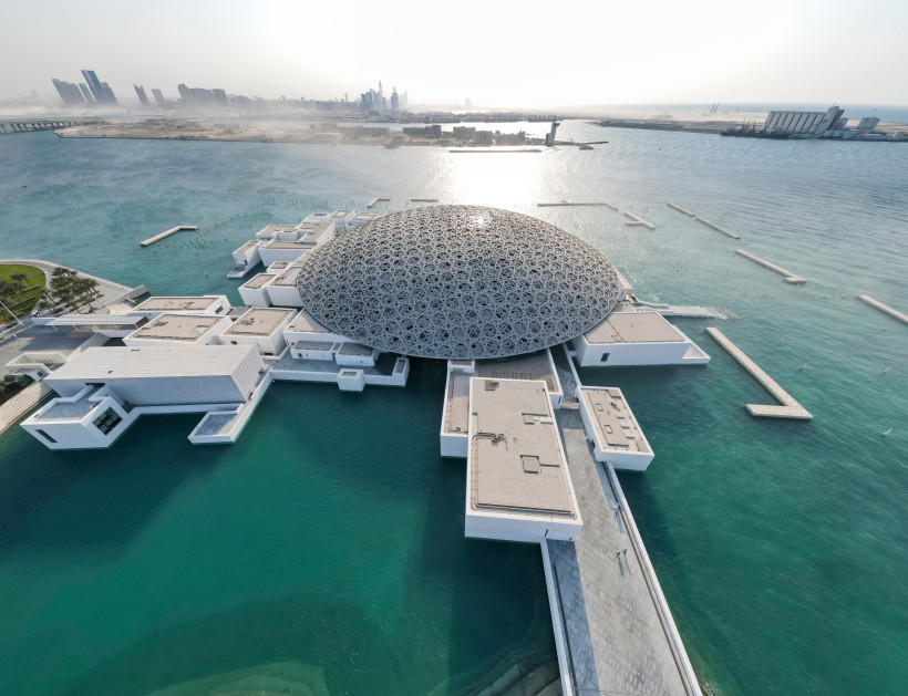 Luftaufnahme des Louvre Abu Dhabi mit seiner ikonischen Kuppel, die auf dem Wasser zu schweben scheint, mit Blick auf die Skyline von Abu Dhabi im Hintergrund