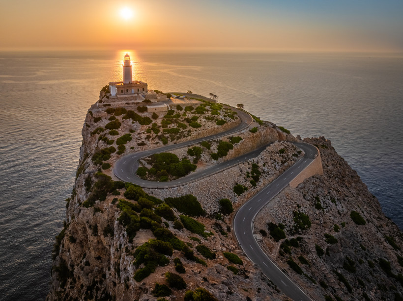 Leuchtturm am Cap de Formentor auf einer Felsklippe mit Serpentinenstraße, im Hintergrund das Meer bei Sonnenuntergang