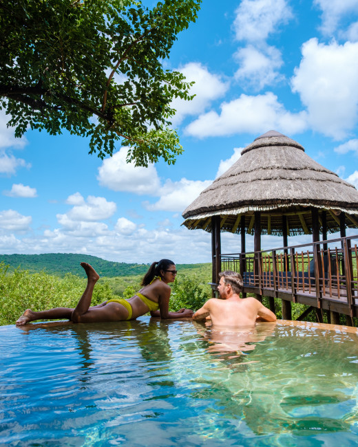 Südafrika Ein Paar entspannt gemeinsam in einem Infinity-Pool mit Blick ins grüne Buschland. Die Frau liegt auf dem Bauch, der Mann lehnt am Beckenrand. Im Hintergrund steht eine Hütte mit Strohdach, darüber ein blauer Himmel mit weißen Wolken.