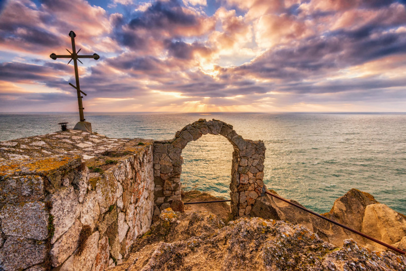 Steinernes Tor und Metallkreuz auf den Klippen von Kap Kaliakra, Bulgarien. Im Hintergrund das Schwarze Meer bei dramatischem Sonnenuntergang mit strahlendem Licht zwischen den Wolken.
