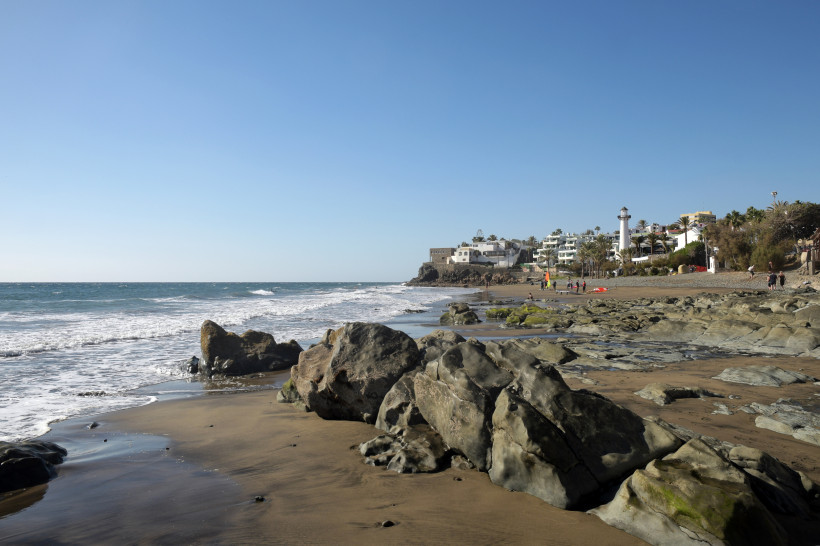Felsiger Strand Playa del Águila bei San Agustín mit Brandung und Küstenbebauung im Hintergrund