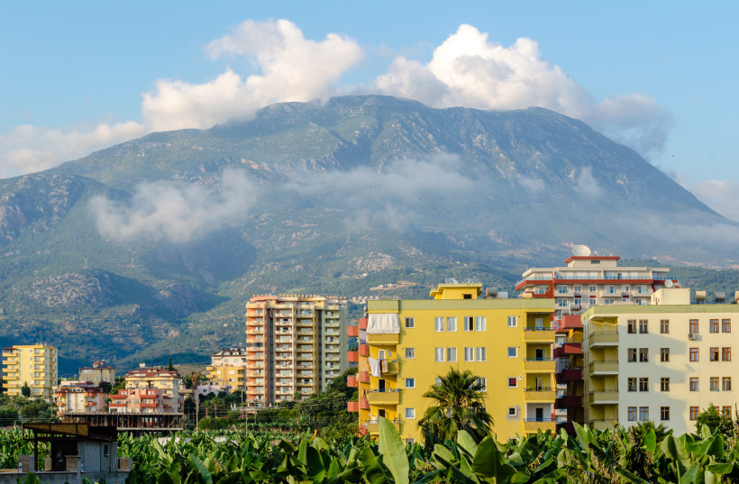 Das Bild zeigt mehrstöckige Wohnhäuser in hellen Farben am Rand der Stadt Alanya. Im Vordergrund befinden sich Bananenplantagen. Im Hintergrund erhebt sich das Taurusgebirge mit bewaldeten Hängen und tiefhängenden Wolken.