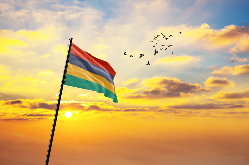 Dieses Bild zeigt die Flagge von Mauritius, die stolz im Wind weht, während die Sonne am Horizont untergeht. Der Himmel ist in warmen Gelb-, Orange- und Blautönen gefärbt, mit verstreuten, weichen Wolken, die von der Abendsonne beleuchtet werden. In der r