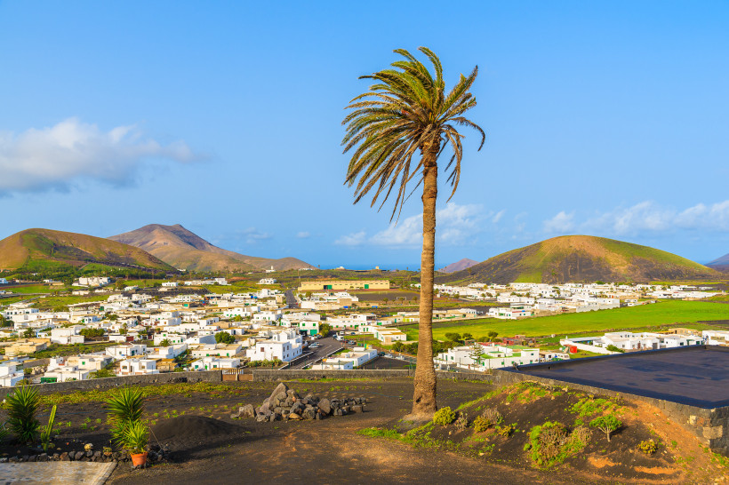 Yaiza auf Lanzarote – Panoramablick über das weiße Dorf im Westen der Insel Panoramablick über Yaiza im Westen von Lanzarote, mit einer einzelnen Palme im Vordergrund, weißen Häusern und den umliegenden Vulkanbergen.
