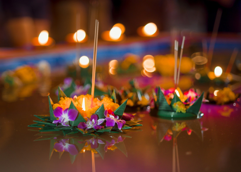 Thailand - Koh Samui  Das Bild zeigt kunstvoll gebastelte schwimmende Gestecke (Krathongs) aus Bananenblättern, bunten Blumen, Kerzen und Räucherstäbchen, die auf einer Wasseroberfläche treiben. Die brennenden Kerzen spiegeln sich sanft im Wasser und verbreiten ein warmes, fe