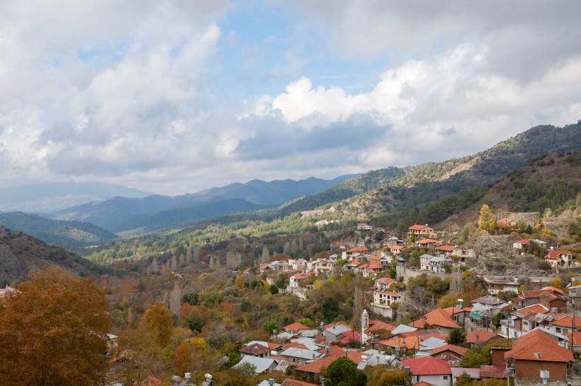 Panoramablick auf ein traditionelles Bergdorf im Troodos-Gebirge mit roten Ziegeldächern, bewaldeten Hängen und Wolken über den Bergen.