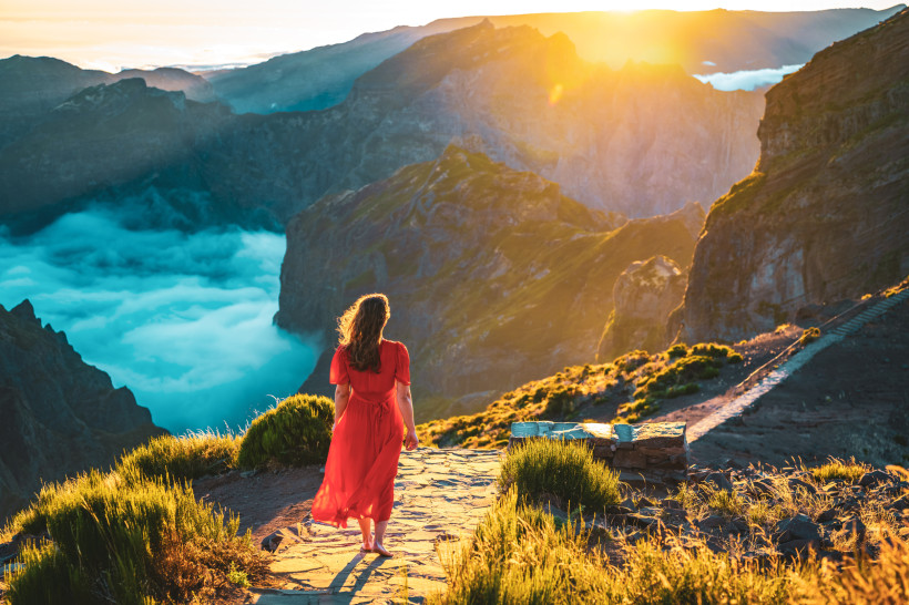 Frau im roten Kleid steht barfuß auf einem Bergpfad auf Madeira und blickt in ein dramatisches Tal, in dem sich Wolken unter ihr sammeln. Die Szene ist in goldenes Morgenlicht getaucht, umgeben von steilen Felswänden und grünen Berghängen