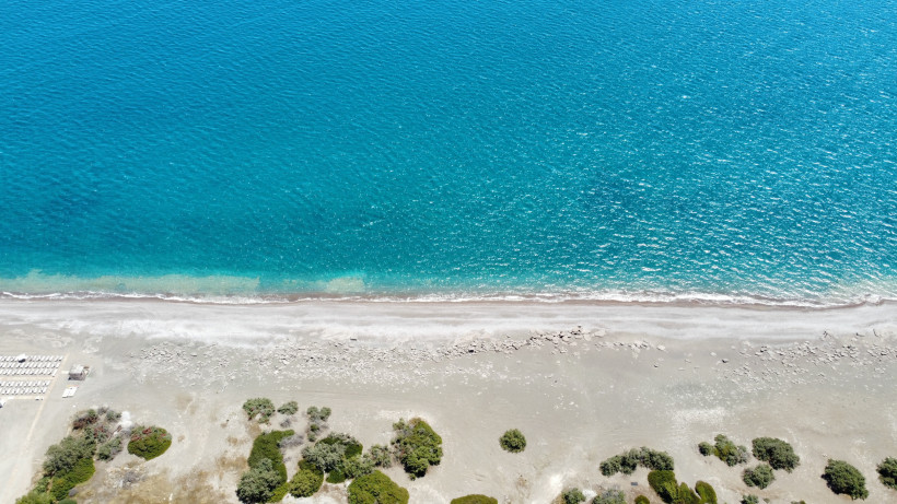 Luftaufnahme eines hellen Sandstrandes mit vereinzelten Steinen, türkisblauem Meer und sanften Wellen aus der Vogelperspektive