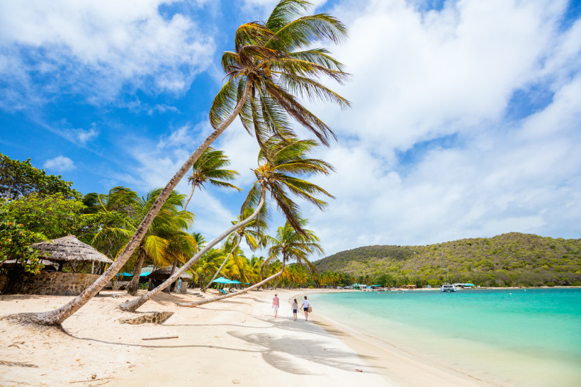 Barbados Breiter Sandstrand mit sanft geschwungenen Palmen, die sich in den blauen Himmel neigen. Drei Menschen spazieren entspannt am türkisfarbenen Meer entlang. Im Hintergrund liegen grüne Hügel und einige Boote vor Anker. Der Himmel ist leicht bewölkt und stra
