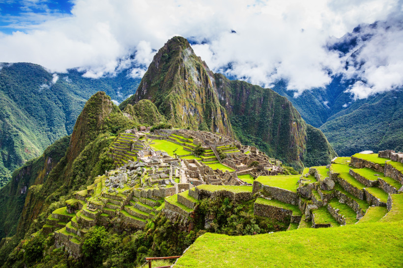 Peru Panoramablick auf die Ruinen von Machu Picchu in Peru, umgeben von üppig grünen Andenbergen. Die steinernen Terrassen und Bauwerke der Inka-Stadt liegen auf einem Bergrücken, im Hintergrund ragt ein spitzer, bewaldeter Berg auf. Der Himmel ist leicht bewö