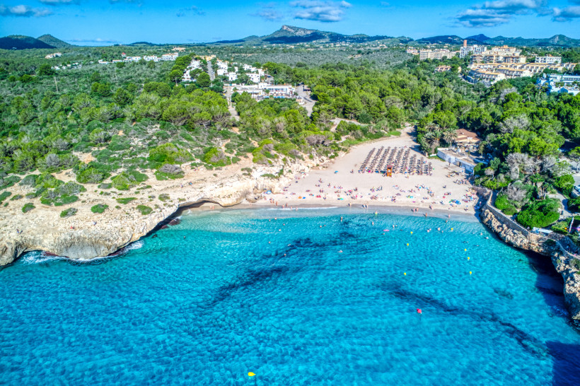 Drohnenaufnahme der Cala Domingos Gran in Calas de Mallorca mit breitem Sandstrand, türkisfarbenem Wasser und Hotels auf den Felsplateaus