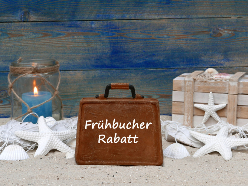 Koffer mit der Aufschrift „Frühbucher Rabatt“ im Sand mit Muscheln und Kerzen – Symbolbild für Frühbucher-Angebote und günstige Urlaubsreisen