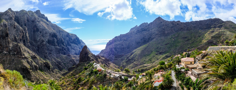 Dramatisches Panorama des Masca-Tals auf Teneriffa Weitwinkelaufnahme des grünen Masca-Tals auf Teneriffa, eingefasst von steil aufragenden dunklen Felswänden. In der Bildmitte verläuft ein kleiner Weg durch das Gebirge Richtung Ozean. Vereinzelte Häuser mit roten Dächern und Palmen sind auf den Hängen zu