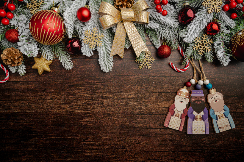 Oben im Bild hängt ein weihnachtlicher Kranz mit verschneiten Tannenzweigen, roten Kugeln, Beeren, goldenen Schneeflocken und einer großen goldenen Schleife. Unten rechts liegen drei weihnachtliche Holzfiguren der Heiligen Drei Könige auf einer dunklen Ho