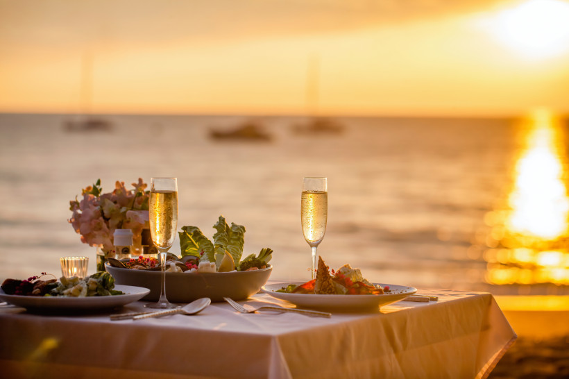 Romantisches Abendessen am Strand mit Blick auf den Sonnenuntergang Elegant gedeckter Tisch mit Champagnergläsern und Speisen direkt am Strand bei goldenem Sonnenuntergang