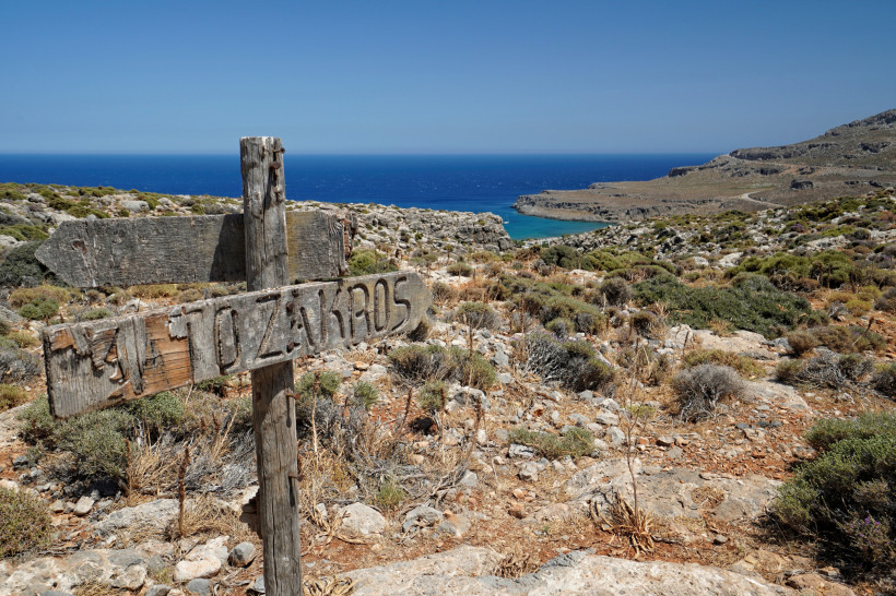 Verwittertes Holzschild mit der Aufschrift 'Zakros' in einer felsigen Landschaft mit Blick auf das blaue Meer auf Kreta