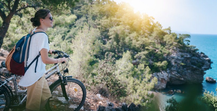 Eine Frau mit Rucksack steht mit ihrem Fahrrad an einem Aussichtspunkt im Wald. Sie blickt lächelnd auf das Meer und eine bewaldete Felsenküste. Die Sonne scheint durch die Bäume, es herrscht eine entspannte, sommerliche Atmosphäre.