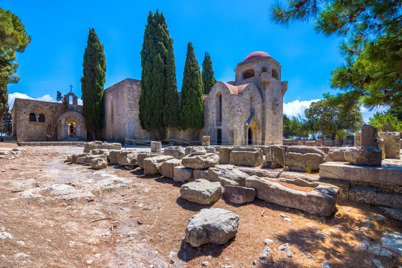 Historische byzantinische Kirche auf Rhodos mit Zypressen, roter Kuppel und antiken Steinruinen im Vordergrund