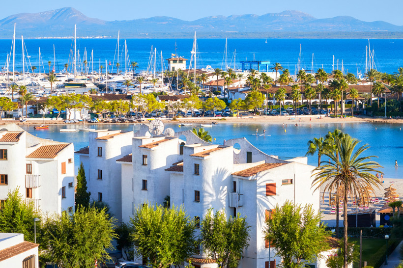 Blick auf den Yachthafen von Port d’Alcúdia mit weißen Gebäuden, Strand und Palmen