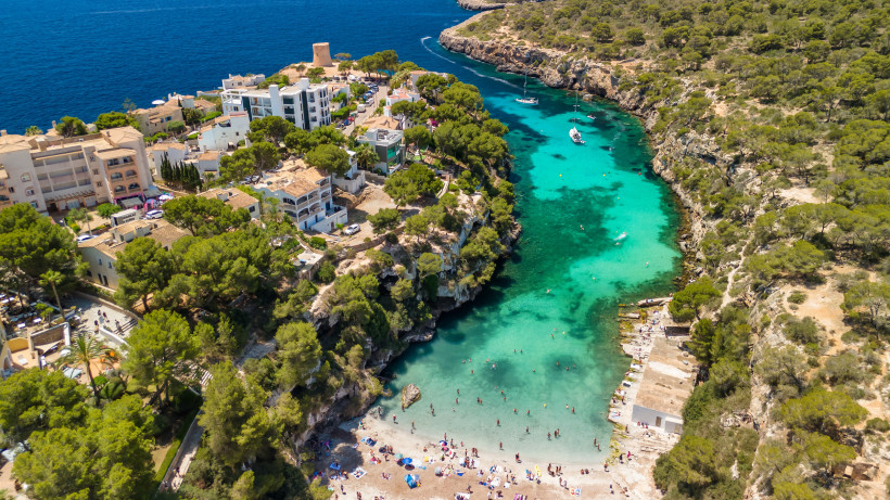 Mallorca - Cala Pi Eine atemberaubende Bucht mit türkisblauem Wasser auf Mallorca, umgeben von steilen Felsklippen mit üppigen Pinienwäldern. Kleine Boote liegen vor Anker, während Menschen an einem hellen Sandstrand baden und sonnen. Im Hintergrund sieht man eine Siedlung