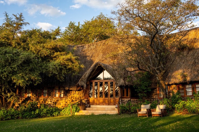 Kenia Gemütliche Lodge mit Natursteinmauern, großen Fenstern und traditionellem Reetdach, umgeben von Bäumen und Büschen. Vor dem Eingang stehen zwei Holzstühle auf gepflegtem Rasen, goldenes Licht fällt auf die Szene