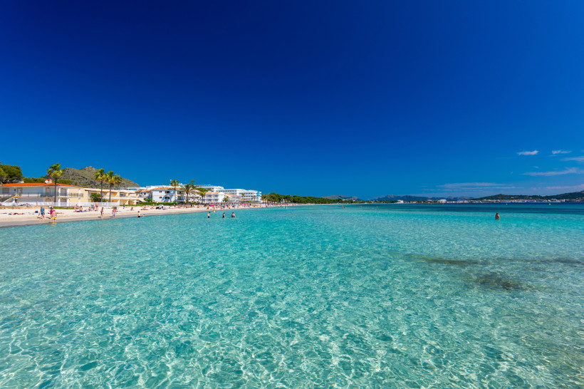 Playa de Muro mit flach abfallendem Strand und seichtem, klarem Wasser, im Hintergrund Gebäude und Palmen