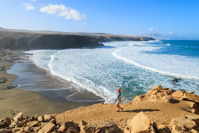 Wanderer mit rotem Hut auf Felsen über der Küste von La Pared auf Fuerteventura, Blick auf den wilden Atlantik mit hohen Wellen