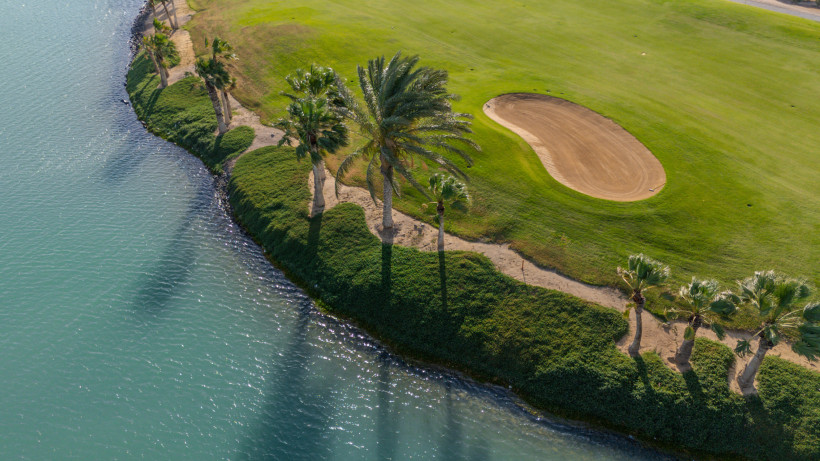 Ägypten - Hurghada Luftaufnahme eines gepflegten Golfplatzes mit grüner Rasenfläche und Palmen, die sich entlang eines ruhigen Gewässers erstrecken. Rechts ist ein Sandbunker sichtbar, links das türkisfarbene Wasser mit leichtem Wellenschlag.