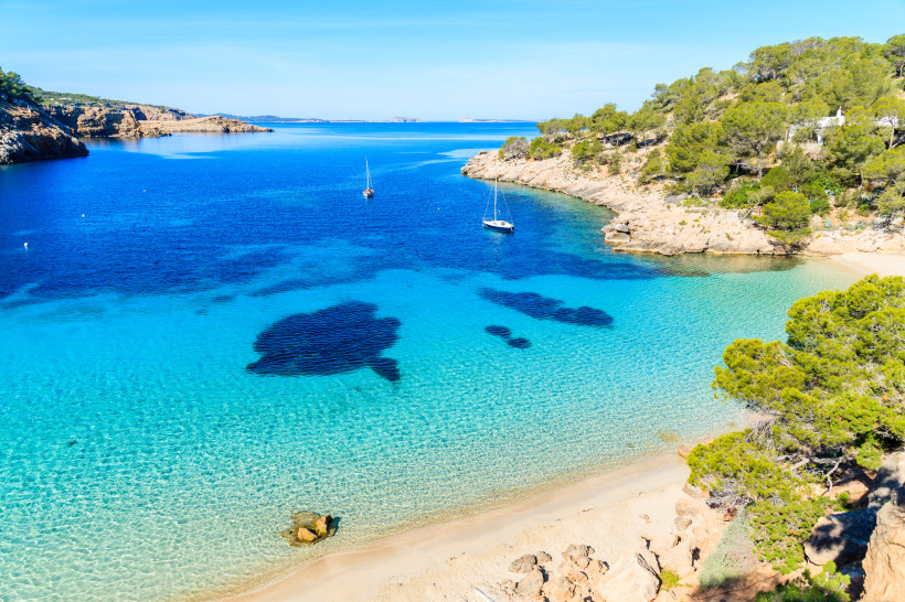 Ibiza - Cala Salada Blick auf die Bucht Cala Saladeta auf Ibiza mit glasklarem türkisblauem Wasser, hellem Sandstrand und bewaldeten Felsen. Zwei Segelboote ankern in der Bucht.