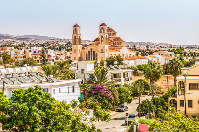 Blick auf die Agioi Anargyroi Kirche in Paphos mit rotbraunen Kuppeln, Palmen und Wohnhäusern im Vordergrund.