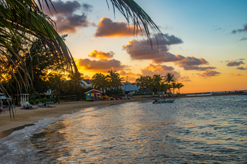 Jamaika Romantischer Sonnenuntergang an einem Strand in Jamaika. Der Himmel ist in warmen Orange- und Violetttönen gefärbt, während Palmen und bunte Strandhütten die Szenerie säumen. Kleine Boote liegen im ruhigen Wasser, einige Menschen entspannen auf Liegen am