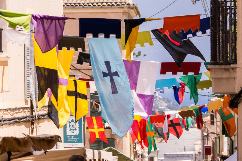 Mallorca Bunte Fahnen und Wimpel hängen quer über einer Altstadtstraße. Sie tragen Kreuze und einfache Muster in leuchtenden Farben wie Gelb, Rot, Blau, Schwarz und Lila. Im Hintergrund sind Häuserfassaden, Fensterläden und ein Firmenschild mit der Aufschrift 'Tor
