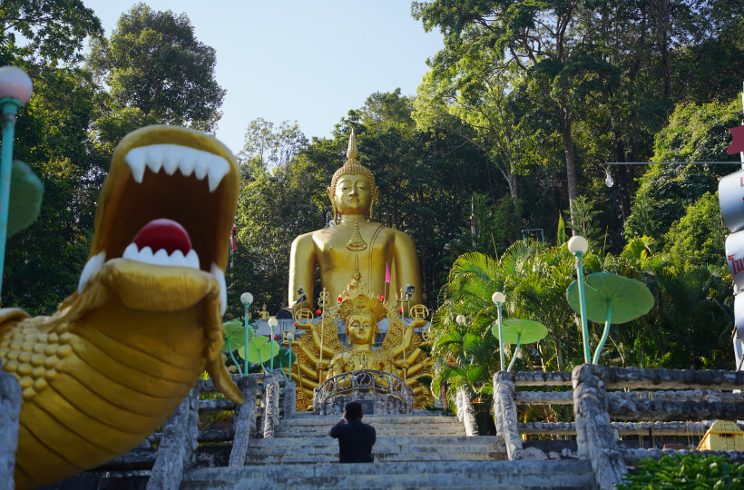 Thailand - Krabi Das Bild zeigt eine imposante goldene Buddha-Statue, die auf einem von Palmen und Dschungelvegetation umgebenen Hügel thront. Im Vordergrund führt eine breite Treppe mit Geländern aus Stein zu der Statue hinauf. Links ist eine große, goldene Drachenfigur