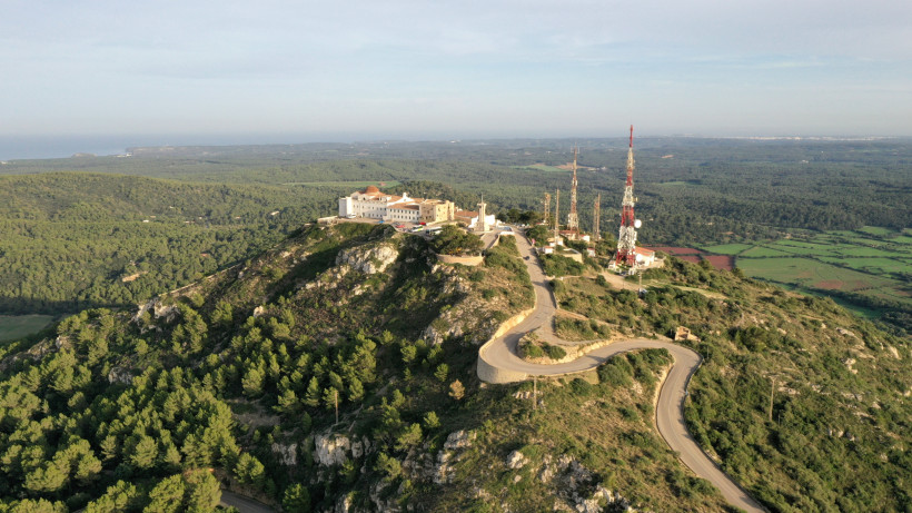Menorca - Monte Toro Luftaufnahme des Monte Toro, des höchsten Berges Menorcas, mit Klosteranlage, Sendemasten und einer kurvenreichen Straße, umgeben von grünen Wäldern und weiter Landschaft.