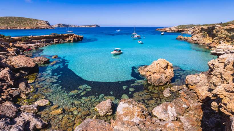 Ibiza - Cala Comte Felsige Bucht Cala Comte auf Ibiza mit kristallklarem, türkisfarbenem Wasser und mehreren Booten, die im ruhigen Meer ankern. Im Vordergrund ragen große Felsen ins Bild, dahinter die weite Küstenlandschaft.
