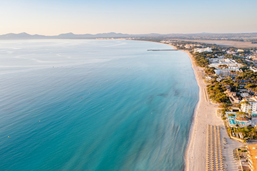 Weitläufige Luftaufnahme der Playa de Alcudia mit langem Sandstrand, ruhiger Wasseroberfläche und Küstenverlauf in Richtung Playa de Muro