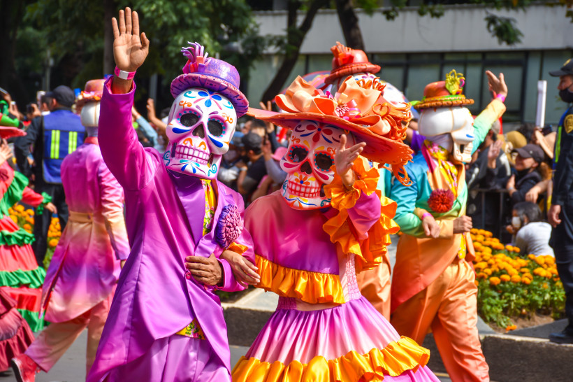 Eine farbenfrohe Parade zum Día de los Muertos (Tag der Toten) mit aufwendig geschmückten Teilnehmern in traditionellen Calavera-Masken und bunten, eleganten Kostümen. Die Feierlichkeiten ehren verstorbene Angehörige und sind ein wichtiger Bestandteil der