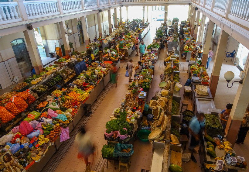 Blick von oben in eine überdachte Markthalle mit zahlreichen bunten Ständen voller frischer Früchte, Gemüse, Kräuter, Gewürze und lokaler Produkte. Menschen schlendern durch die Gänge zwischen den Verkaufsständen.