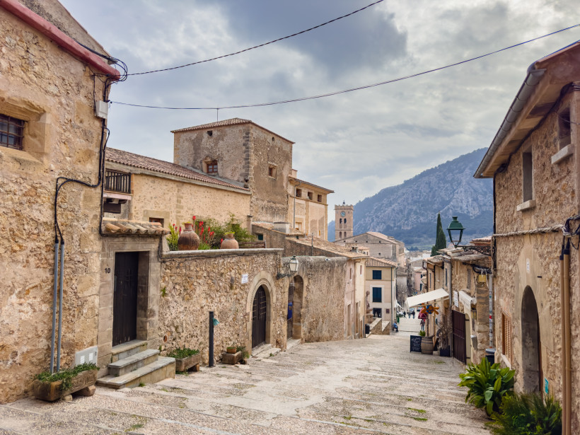 Steile Naturstein-Gasse in der Altstadt von Pollença mit traditionellen Häusern und Blick auf den Kirchturm vor der Kulisse der Serra de Tramuntana.