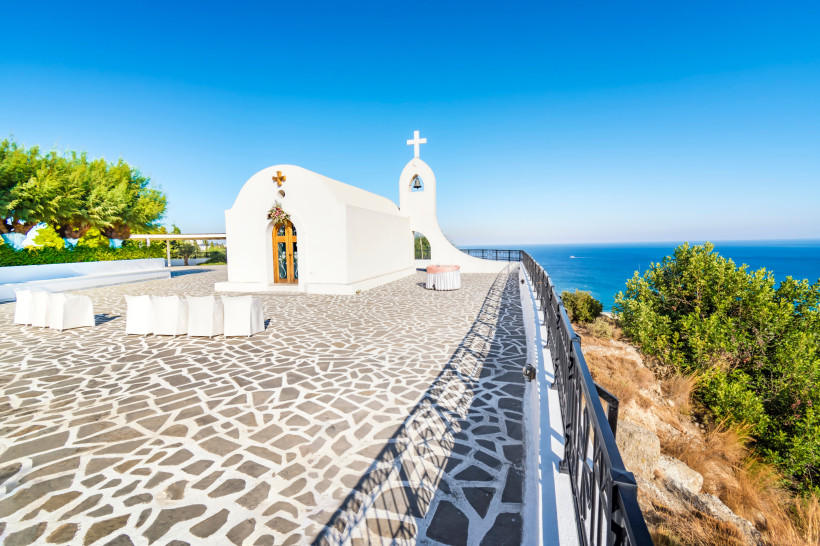 Weiße griechische Kapelle auf einer Klippe mit gepflasterter Terrasse und Blick auf das blaue Meer