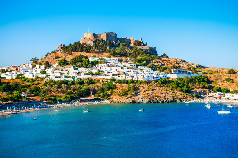 Blick auf Lindos auf Rhodos mit weißem Dorf, Burganlage auf dem Hügel und türkisblauer Badebucht