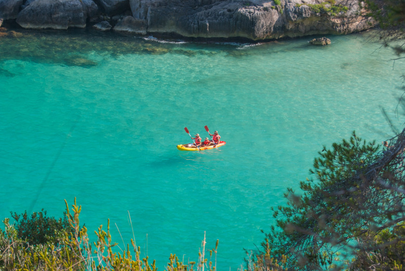 Drei Personen in einem gelben Kajak paddeln auf türkisfarbenem, klarem Wasser entlang einer Felsküste. Alle tragen rote Schwimmwesten und Hüte. Das Bild ist von oben aufgenommen und eingerahmt von grünem Gebüsch und Pinien, die am Bildrand sichtbar sind. 