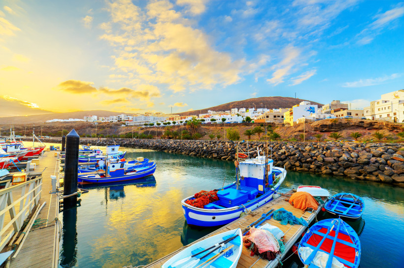 Fuerteventura Kleine bunte Fischerboote liegen in einem ruhigen Hafenbecken vor einer Küstenstadt mit weißen Häusern. Die Abendsonne taucht Himmel und Wasser in warmes Licht
