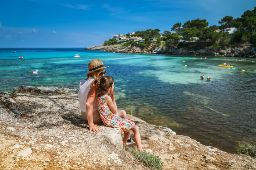 Mallorca Eine Frau mit Strohhut sitzt mit einem kleinen Mädchen auf einem Felsen am Ufer und blickt aufs türkisfarbene Meer. Beide tragen Sommerkleidung, das Kind ein bunt geblümtes Kleid. Im Hintergrund ist eine geschützte Bucht mit Schwimmern, Tretbooten und Fer