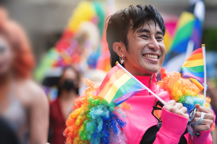 Thailand - Krabi Das Bild zeigt eine lächelnde Person bei einer LGBTQ  Pride-Parade. Sie trägt ein auffälliges Outfit mit pinker Jacke, Regenbogenperücke und herzförmigem Ohrring. In beiden Händen hält sie kleine Regenbogenflaggen. Im Hintergrund sind weitere Teilnehmer*i