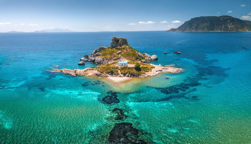 Kos - Kefalos  Drohnenbild der kleinen Insel Kastri vor Kos mit einer weiß-blauen Kapelle, umgeben von türkisfarbenem Wasser. Die Insel ist von Felsen umgeben, im Hintergrund liegt die grüne Küste der Halbinsel.